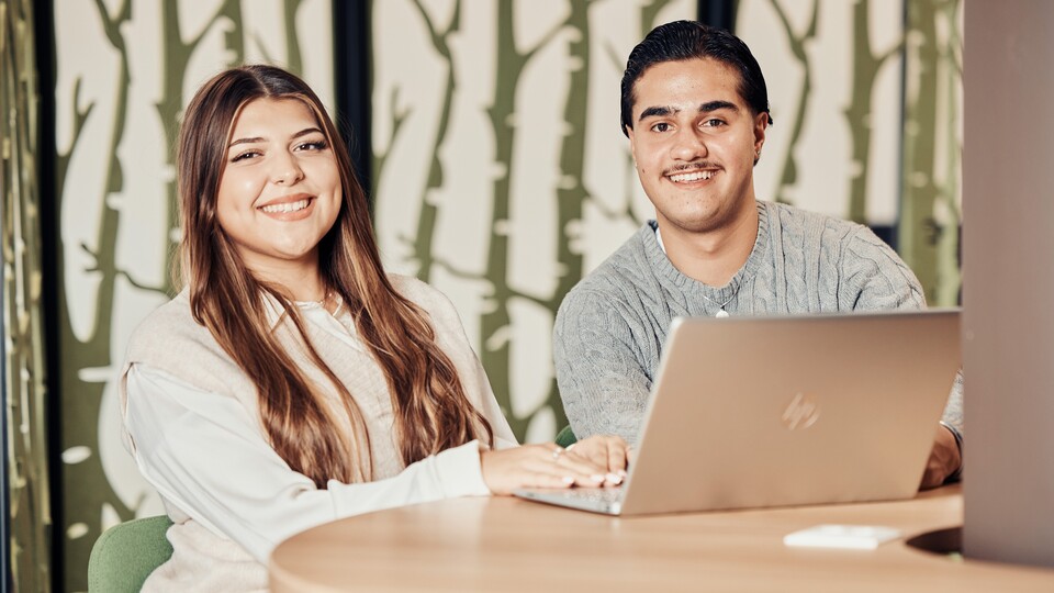 Ein junger Mann und eine junge Frau sitzen an einem hellen Holztisch vor einem Laptop und lachen in die Kamera.