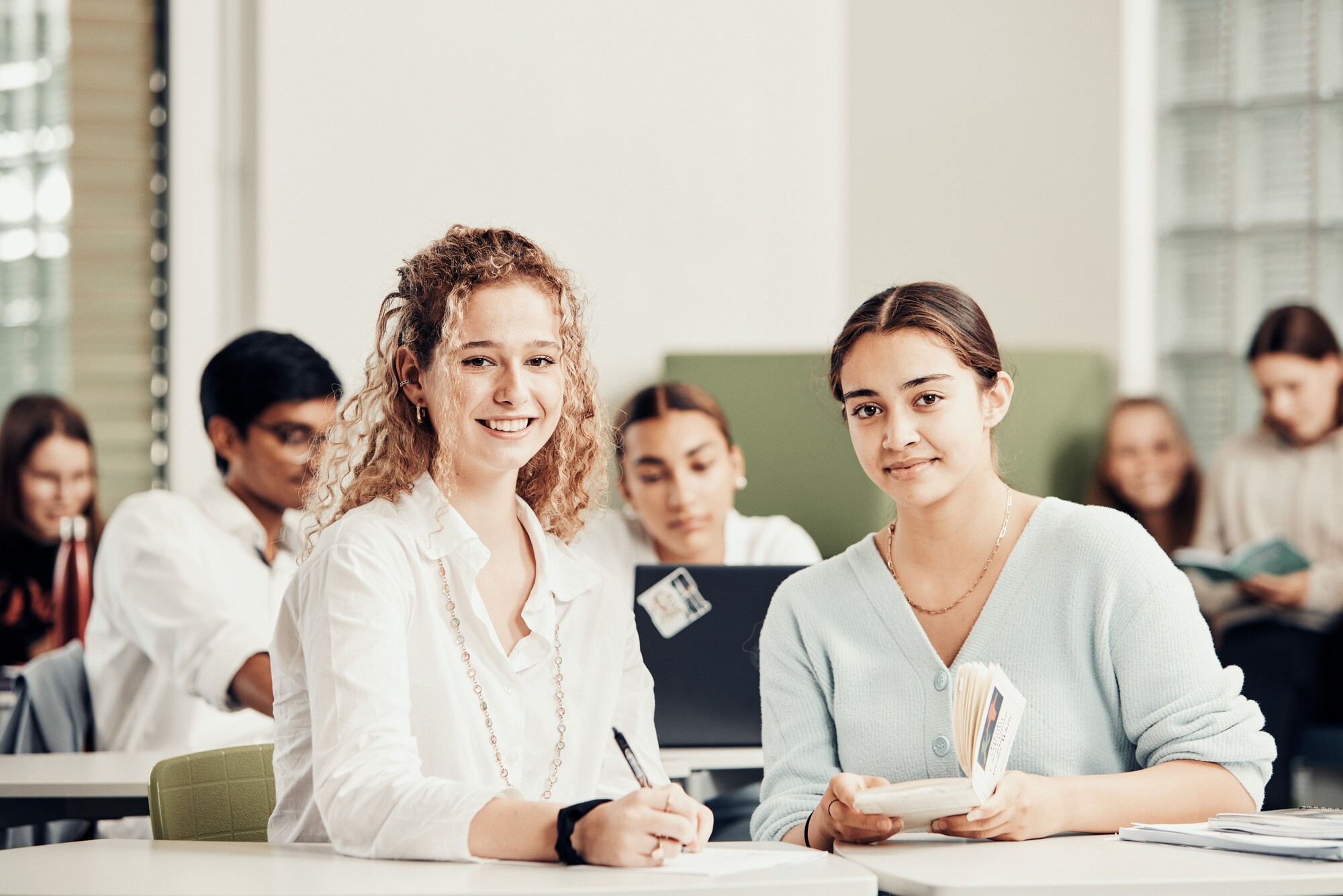 Zwei junge Frauen sitzen im Vordergrund in einem Klassenzimmer, arbeiten mit Buch und Schreiber und lächeln in die Kamera.