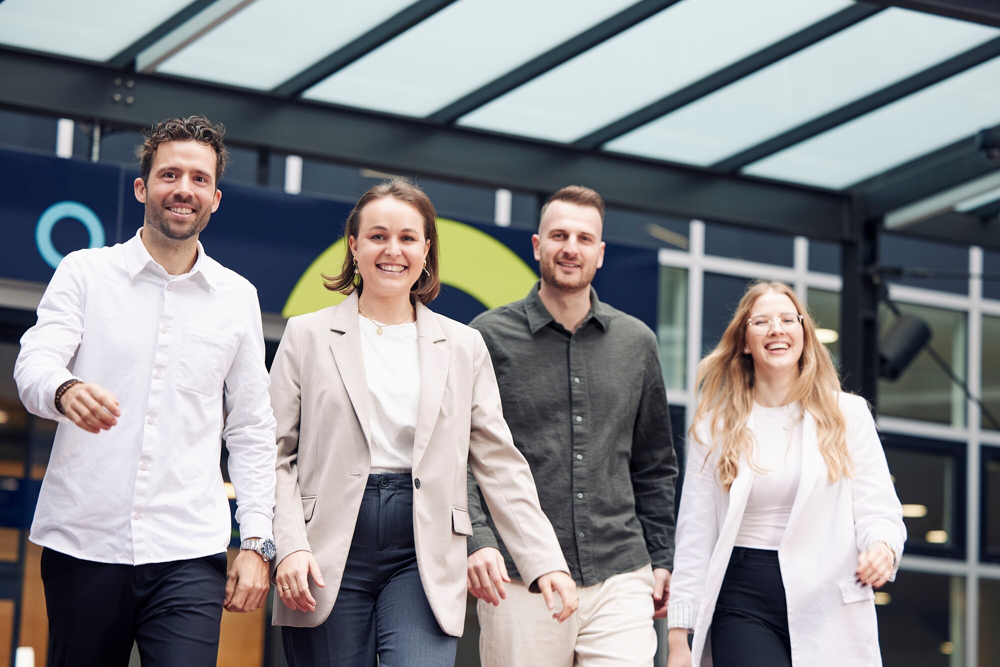 Vier Studierende der KV Luzern Berufsakademie Gut gekleidete Studierende gehend lachend auf die Kamera zu, sie befinden sich vor dem Eingang der KV Luzern Berufsakademie.