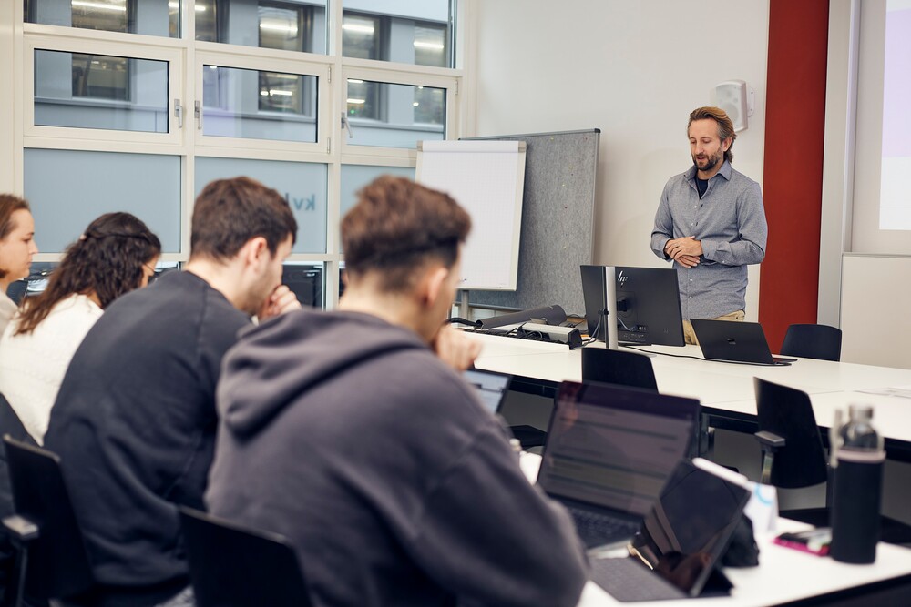 Studierende sitzen aufmerksam an Tischen vor dem Laptop, während ein Dozent an der Tafel spricht.