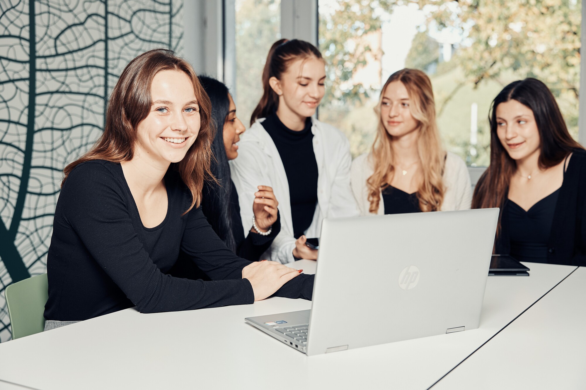 Eine Frauengruppe sitzt an einem Tisch mit Laptop und bespricht etwas, eine der jungen Frauen schaut ins Bild.