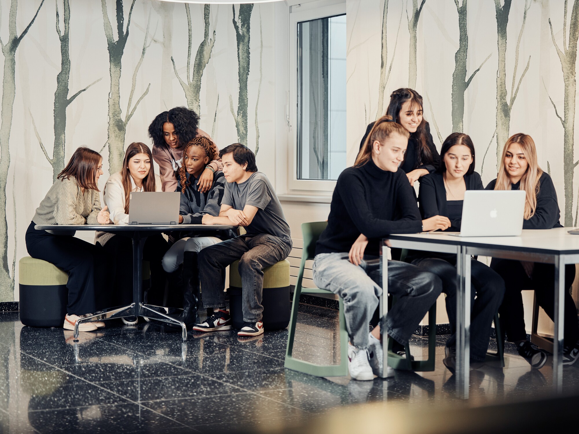 Zwei Gruppen von Lernenden sitzen an zwei separaten Tischen und schauen gemeinsam auf einen Laptop.