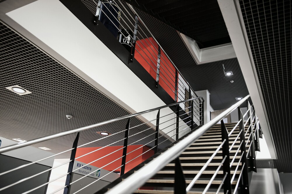 Eine moderne dunkle Treppe im leeren Schulgebäude der KV Luzern mit roten Farbakzenten im Hintergrund.