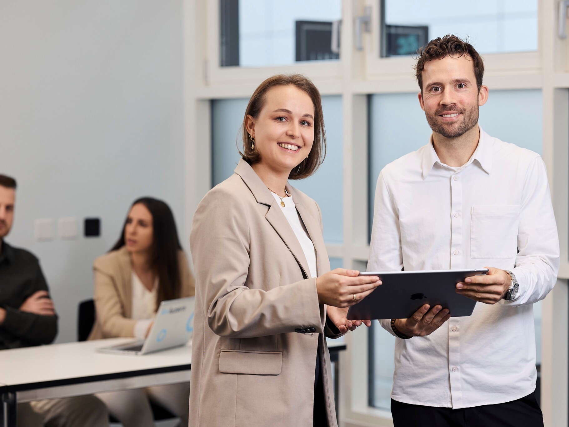 Zwei Studierende der KV Luzern Berufsakademie Ein Mann mit weissem Hemd und eine Frau mit Blazer lächeln in die Kamera, sie stehen vor einem Tisch und halten ein Tablet.