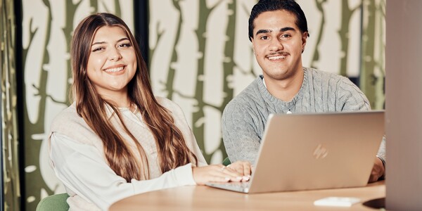 Ein junger Mann und eine junge Frau sitzen an einem hellen Holztisch vor einem Laptop und lachen in die Kamera.