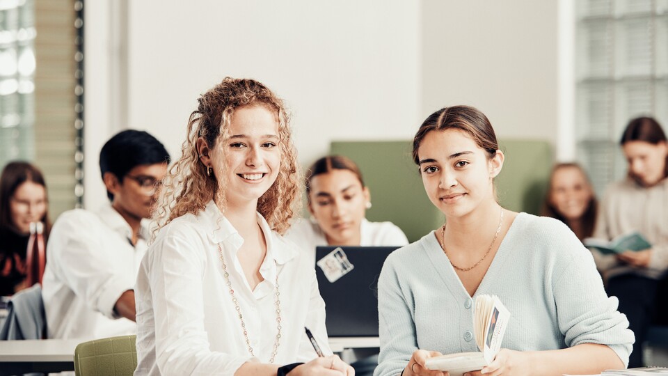 Zwei junge Frauen sitzen im Vordergrund in einem Klassenzimmer, arbeiten mit Buch und Schreiber und lächeln in die Kamera.