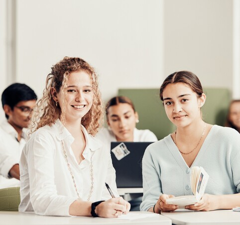 Zwei junge Frauen sitzen im Vordergrund in einem Klassenzimmer, arbeiten mit Buch und Schreiber und lächeln in die Kamera.